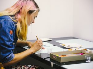 An artist with blonde and pink hair sits at a desk painting.