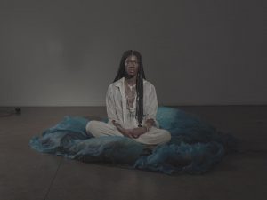 A woman with dark braids dressed in white sits cross legged on a blue sheet on a concrete floor.